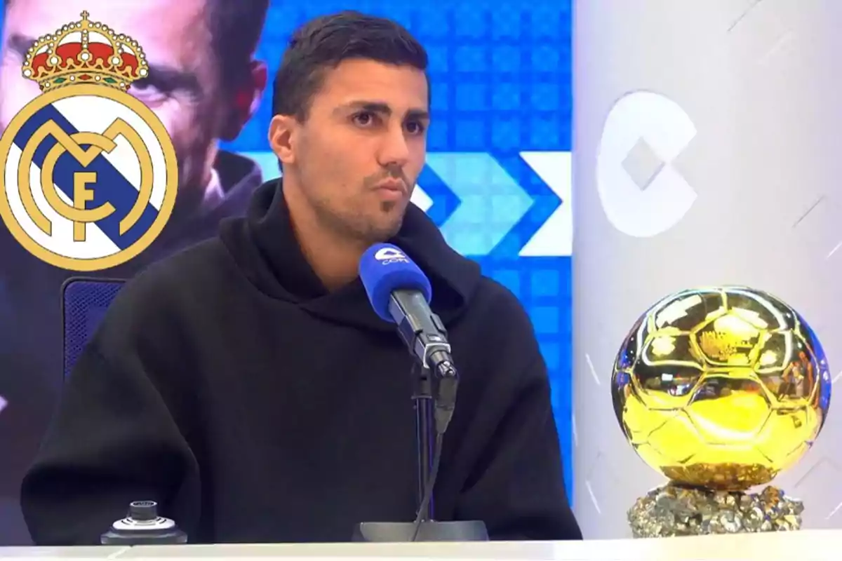 Un hombre en una entrevista con un micrófono azul y un trofeo dorado sobre la mesa, con el logo del Real Madrid en el fondo.