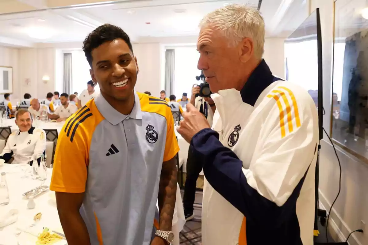 Dos personas con uniformes del Real Madrid conversan y sonríen en un comedor lleno de otros miembros del equipo.