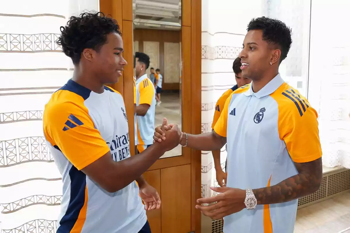 Dos jugadores de fútbol del Real Madrid se saludan con un apretón de manos mientras sonríen, ambos visten uniformes de entrenamiento del equipo.