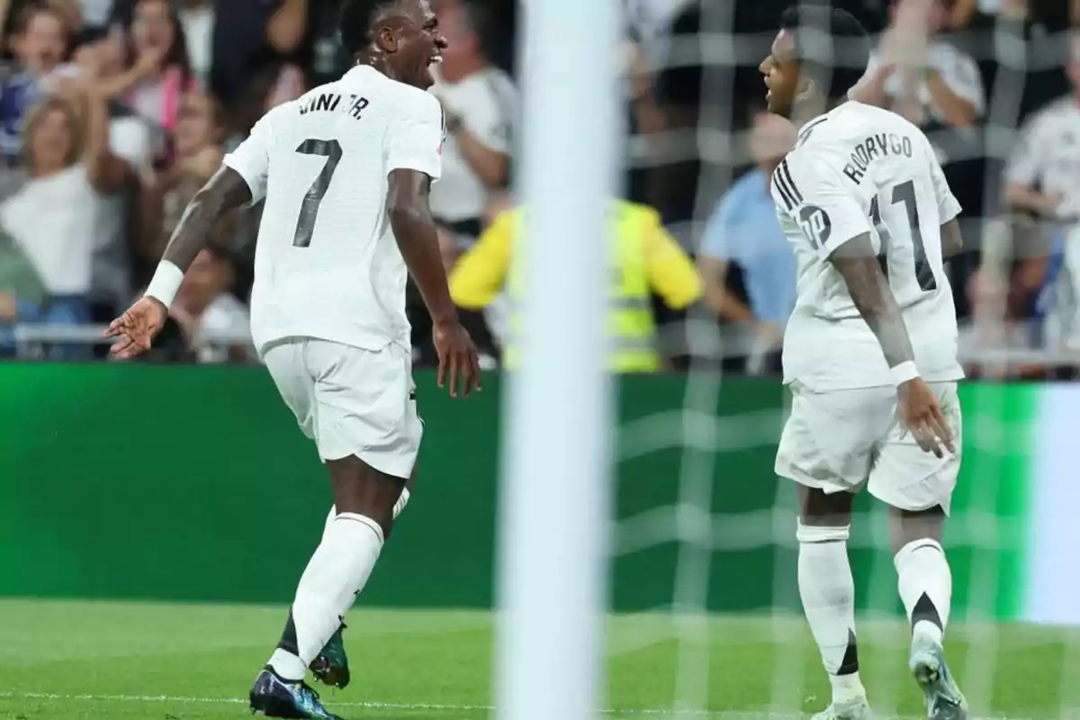 Dos jugadores de fútbol celebran en el campo mientras el público aplaude en el fondo.