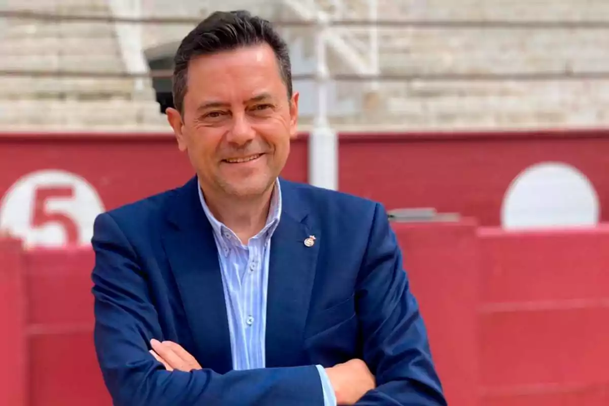 Hombre con traje azul y camisa a rayas, sonriendo y con los brazos cruzados, de pie frente a una pared roja con el número 5 en el fondo.