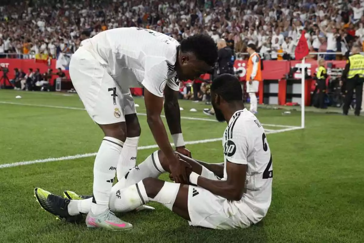Dos jugadores de fútbol del Real Madrid en el campo uno de pie y el otro sentado mientras el público observa. Dos jugadores de fútbol del Real Madrid en el campo uno de pie y el otro sentado mientras el público observa.