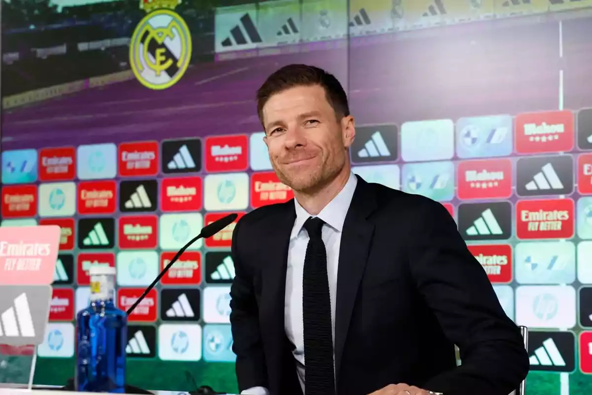 Hombre con traje oscuro y corbata negra sentado en una conferencia de prensa con fondo de logotipos de patrocinadores y el escudo de un equipo de fútbol Hombre con traje oscuro y corbata negra sentado en una conferencia de prensa con fondo de logotipos de patrocinadores y el escudo de un equipo de fútbol
