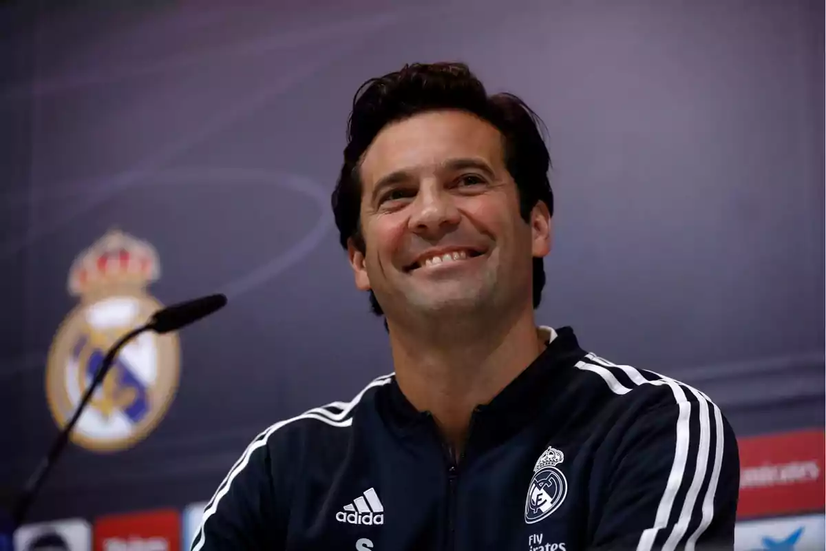 Un hombre sonriente con una chaqueta deportiva del Real Madrid en una conferencia de prensa.