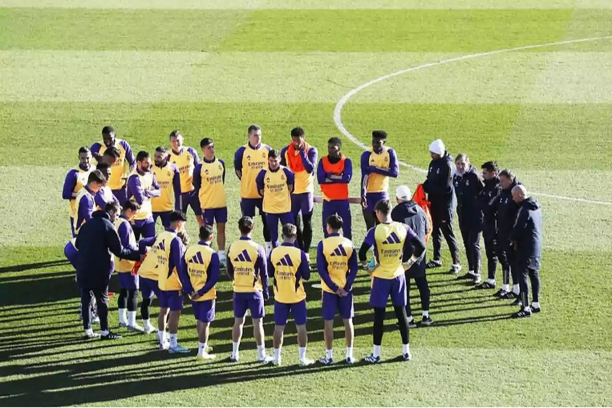 Un grupo de jugadores de fútbol y entrenadores se encuentran reunidos en un campo de entrenamiento, con los jugadores vistiendo chalecos amarillos y uniformes morados.