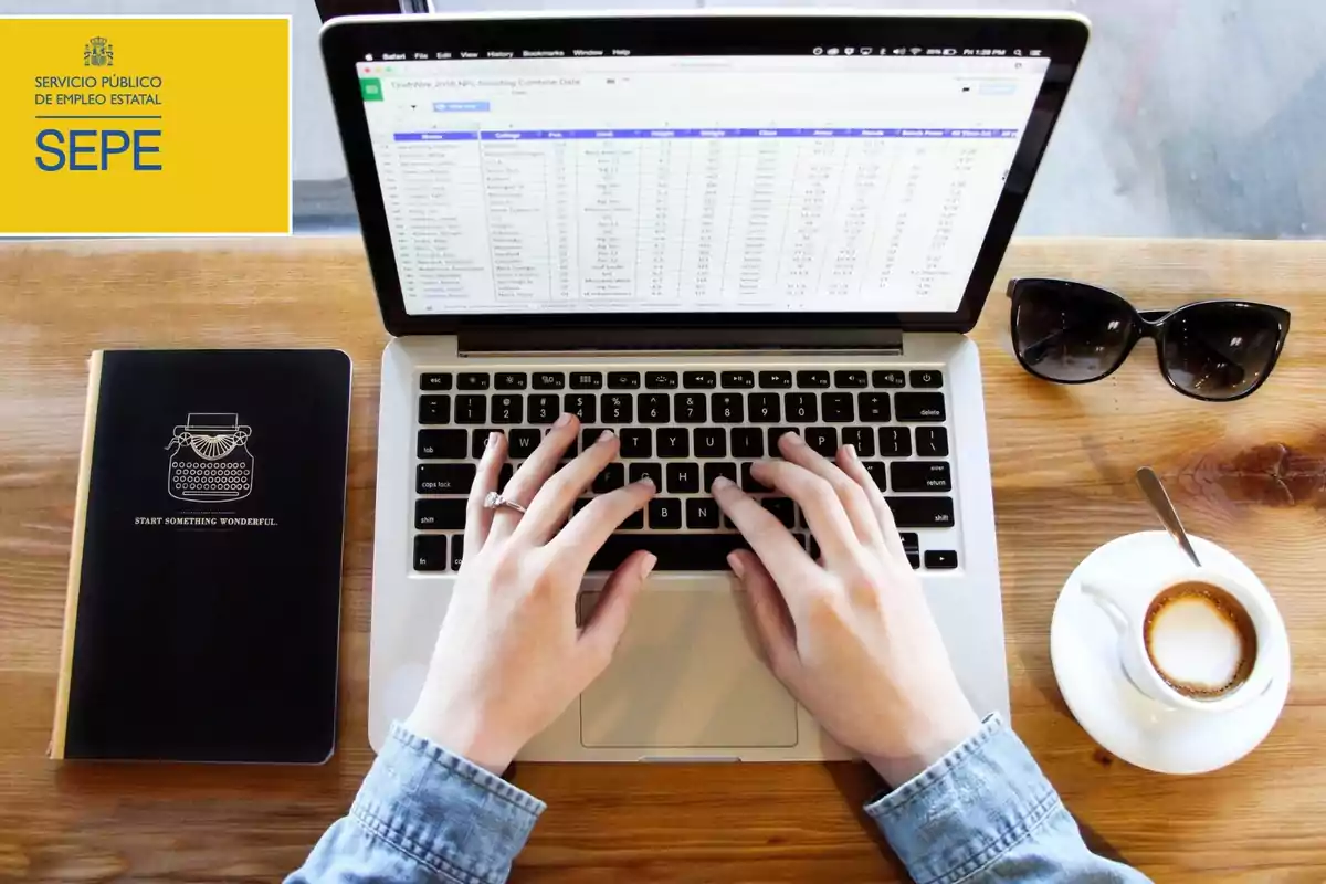 Una persona trabajando en una computadora portátil con una libreta negra, una taza de café y unas gafas de sol sobre una mesa de madera, junto a un logotipo del Servicio Público de Empleo Estatal (SEPE).