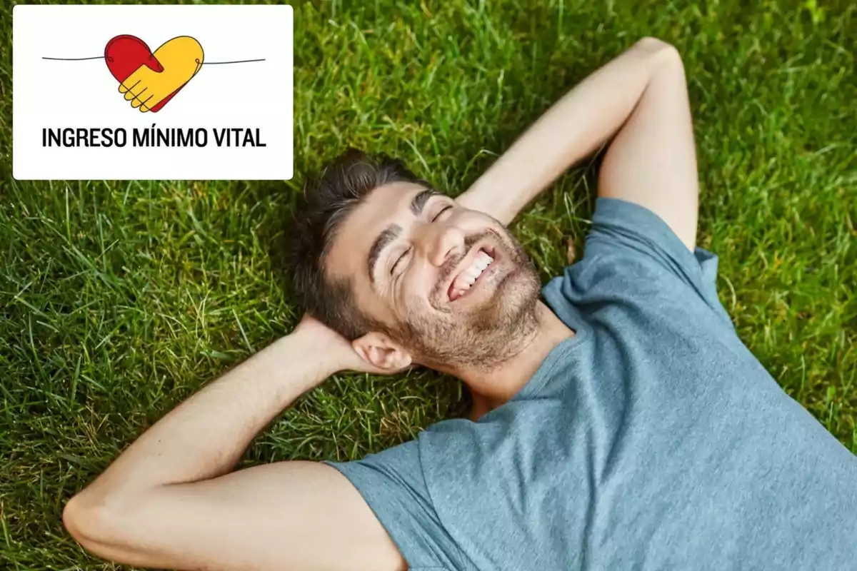 Un hombre sonriente recostado sobre el césped junto a un cartel que dice "Ingreso Mínimo Vital" con un diseño de manos formando un corazón.