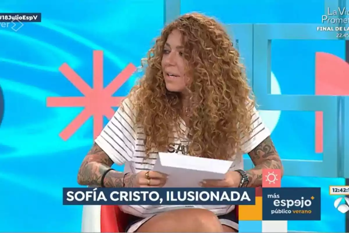 Una mujer con cabello rizado y tatuajes en los brazos está sentada en un programa de televisión, sosteniendo unos papeles y mirando hacia un lado, con un fondo azul y un gráfico rojo detrás de ella. En la parte inferior de la imagen se puede leer el texto Una mujer con cabello rizado y tatuajes en los brazos está sentada en un programa de televisión, sosteniendo unos papeles y mirando hacia un lado, con un fondo azul y un gráfico rojo detrás de ella. En la parte inferior de la imagen se puede leer el texto