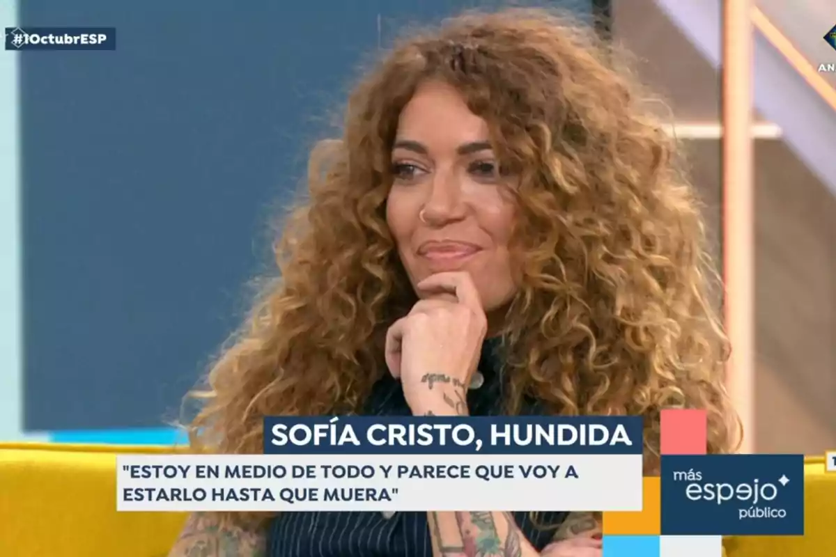 Mujer con cabello rizado y tatuajes en un programa de televisión con el texto 