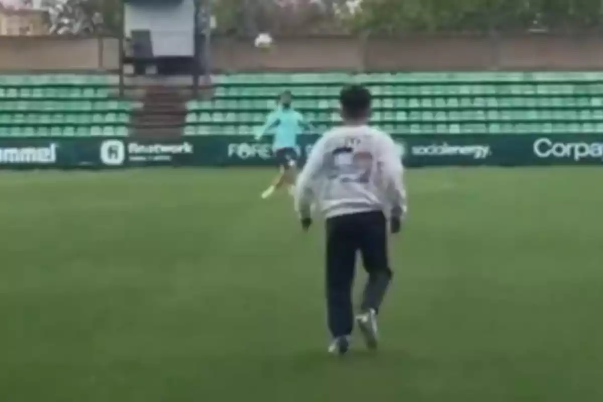 Dos personas jugando al fútbol en un campo con gradas verdes.