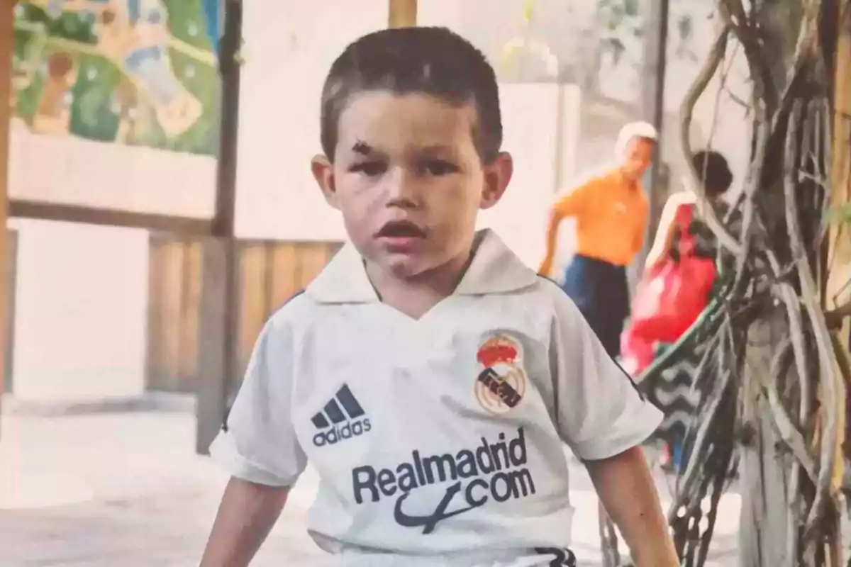 Un niño con una camiseta del Real Madrid aparece en la imagen, con una expresión seria y una cicatriz en la frente, mientras que en el fondo se observan otras personas y un entorno al aire libre.