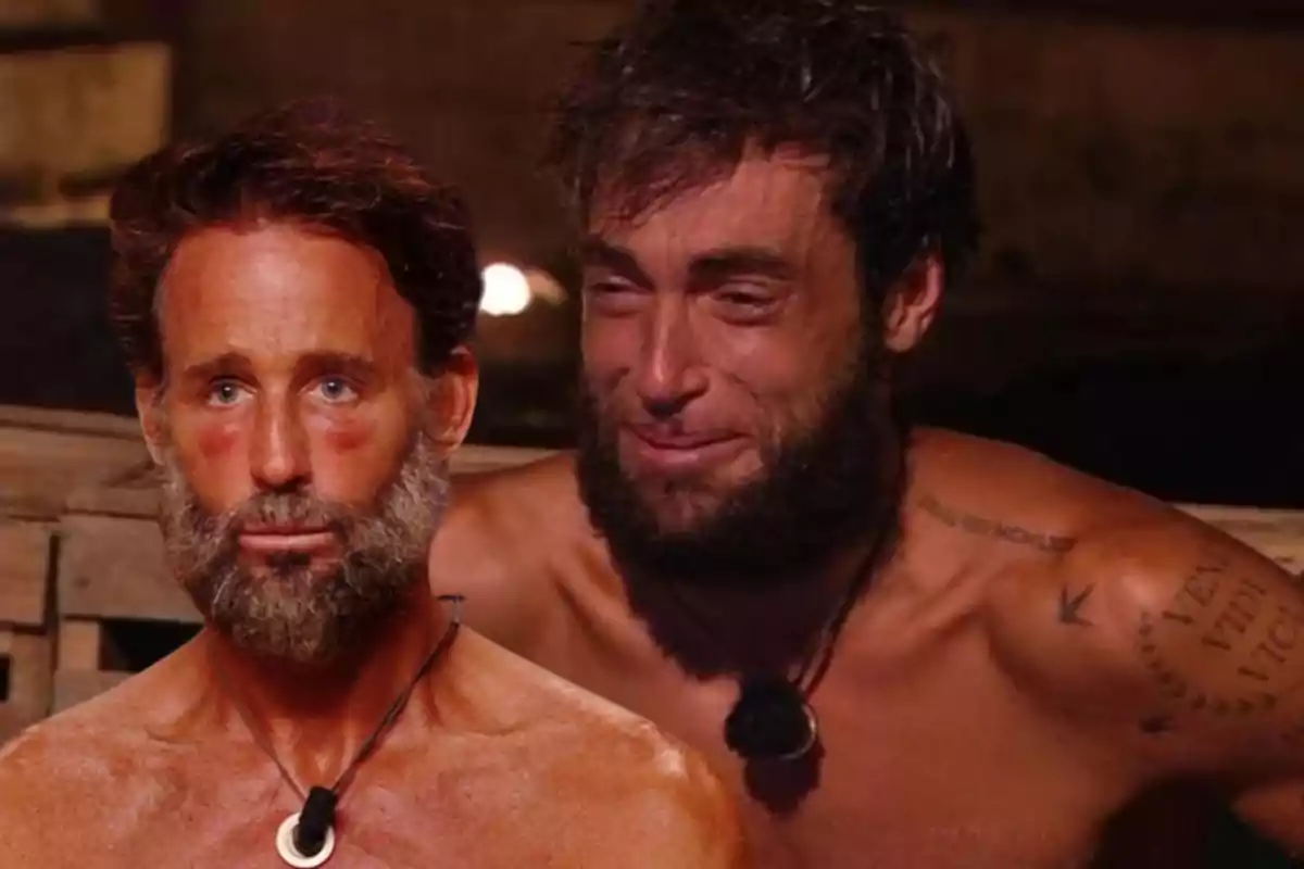 Dos hombres con barba y piel bronceada, uno de ellos con expresión seria y el otro llorando, ambos sin camiseta y usando collares, en un ambiente rústico.