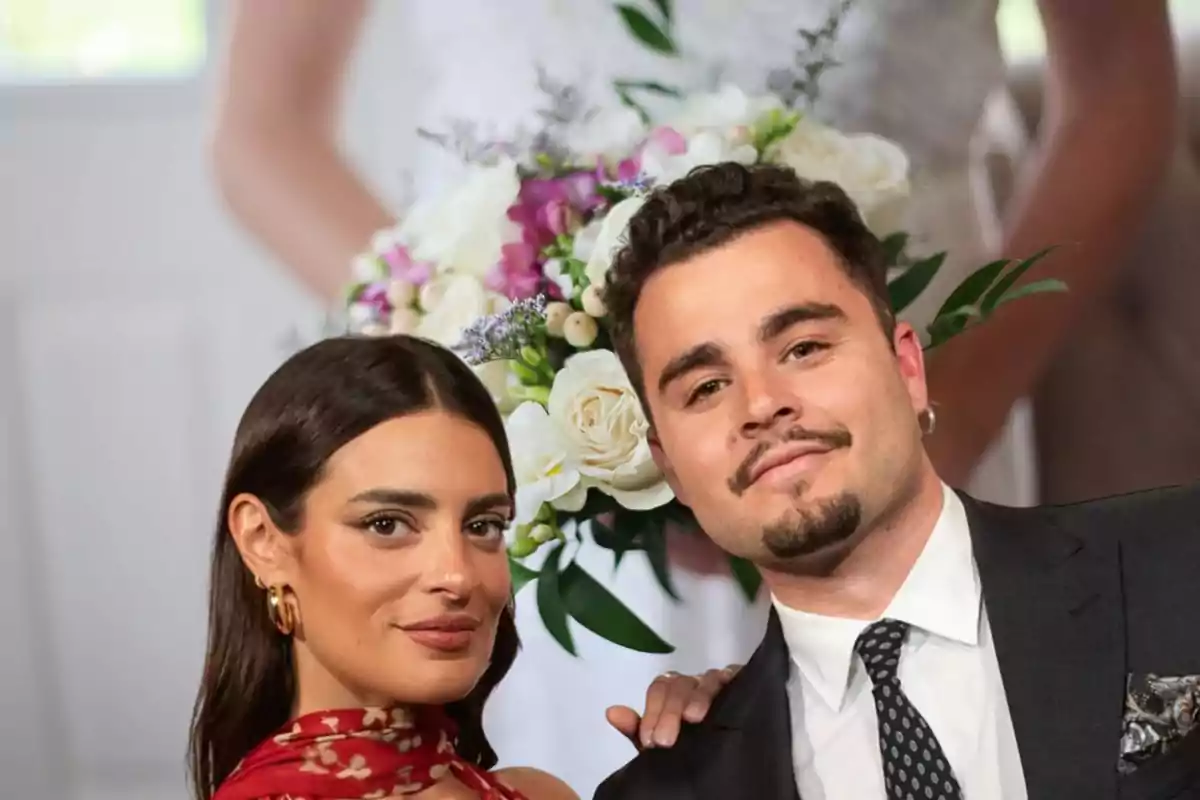 Una mujer y un hombre posan juntos elegantemente vestidos con un ramo de flores al fondo
