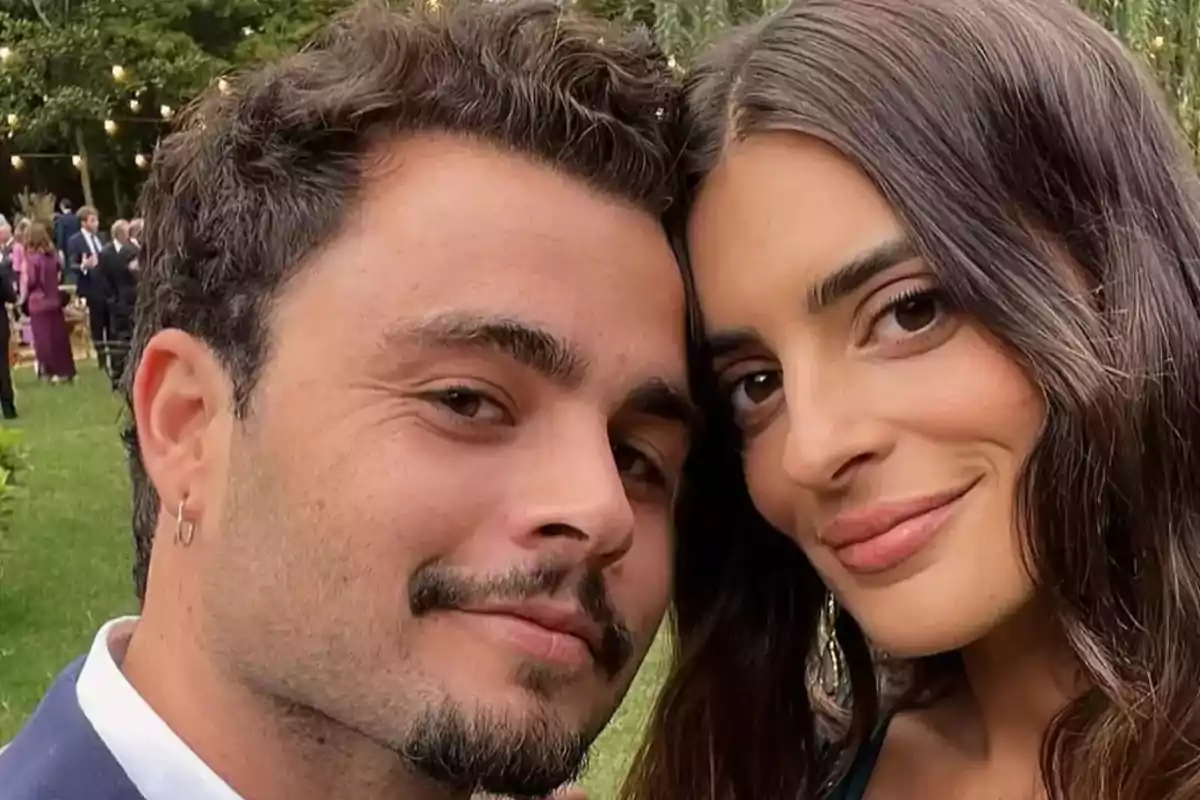 Pareja joven sonríe mientras se toma una selfie en un evento al aire libre con personas al fondo Pareja joven sonríe mientras se toma una selfie en un evento al aire libre con personas al fondo