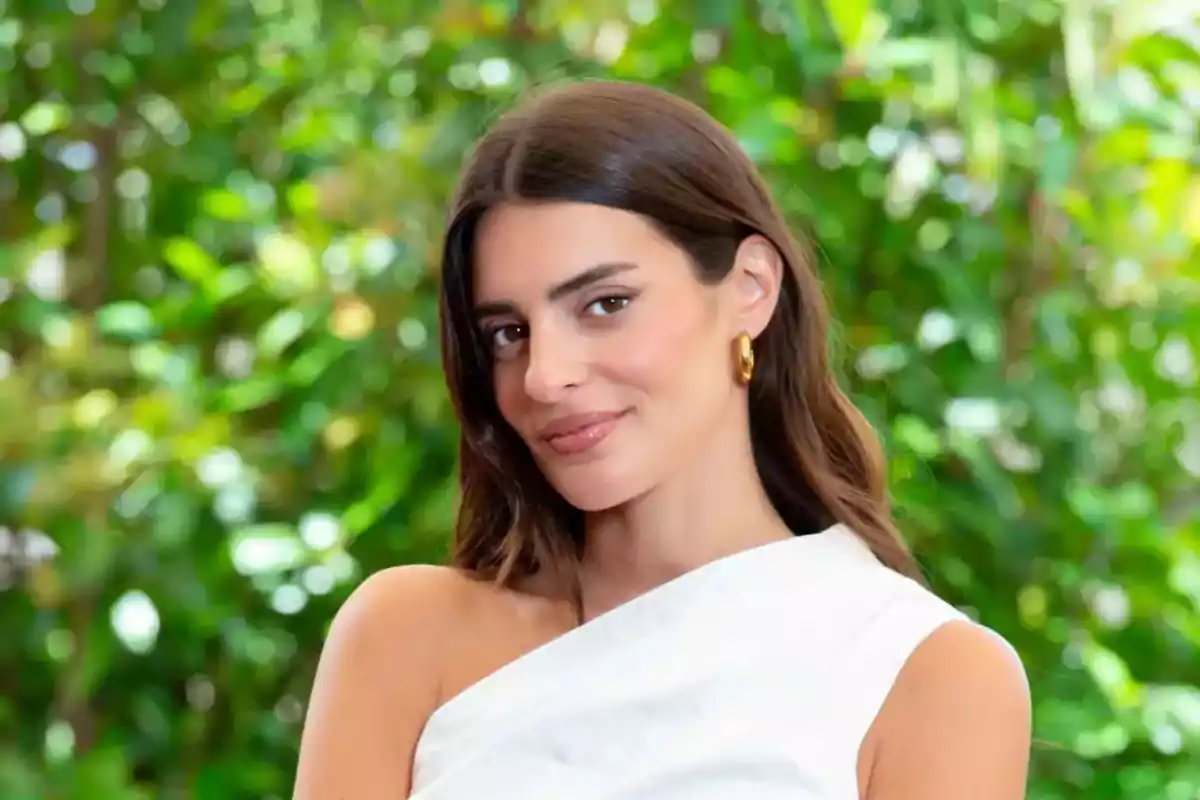 Mujer joven de cabello castaño y liso con vestido blanco de un solo hombro y aretes dorados posando frente a un fondo de vegetación verde Mujer joven de cabello castaño y liso con vestido blanco de un solo hombro y aretes dorados posando frente a un fondo de vegetación verde
