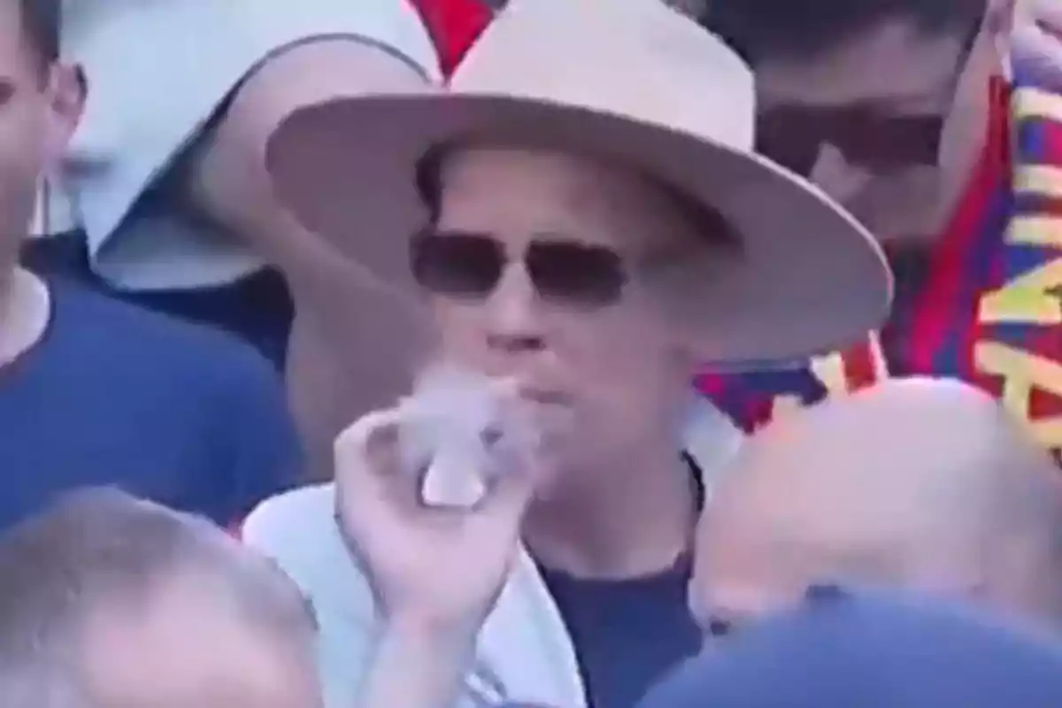 Un hombre con sombrero y gafas de sol está rodeado de personas en un evento mientras sostiene un cigarrillo electrónico. Un hombre con sombrero y gafas de sol está rodeado de personas en un evento mientras sostiene un cigarrillo electrónico.