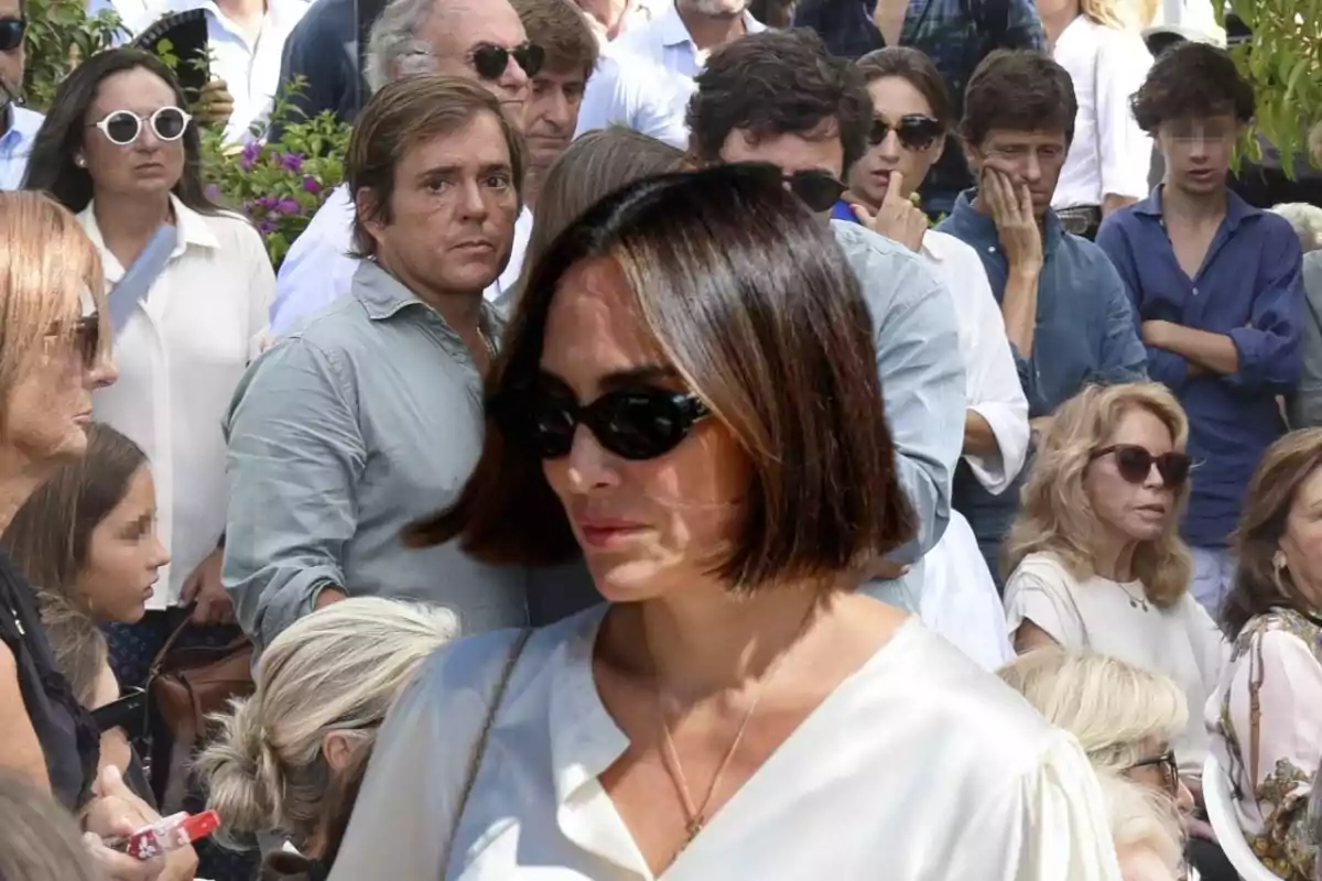 Una mujer con gafas de sol y cabello corto camina entre una multitud de personas, algunas de las cuales también llevan gafas de sol.