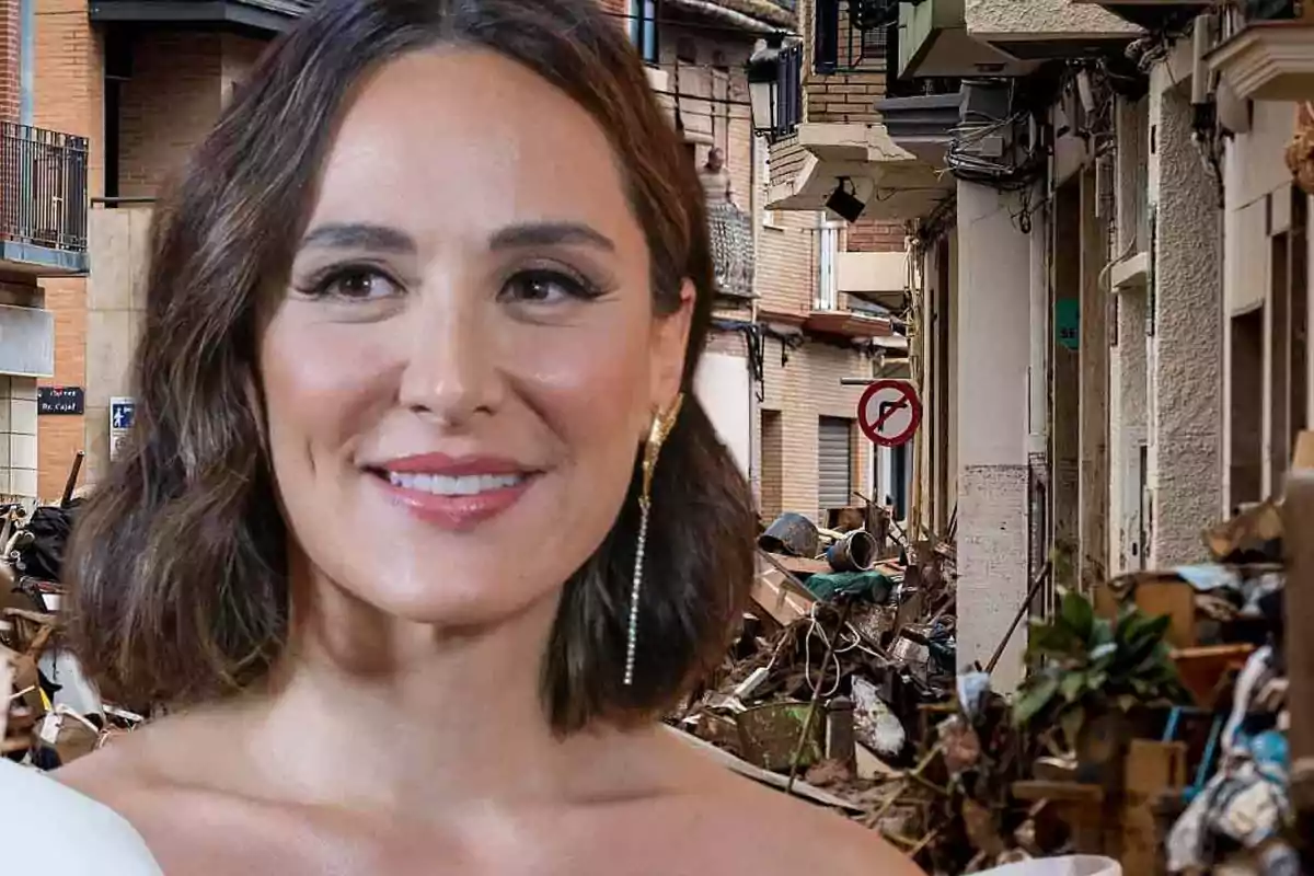 Una mujer sonriente con cabello oscuro y aretes largos está en primer plano, mientras que al fondo se observa una calle con escombros y edificios.