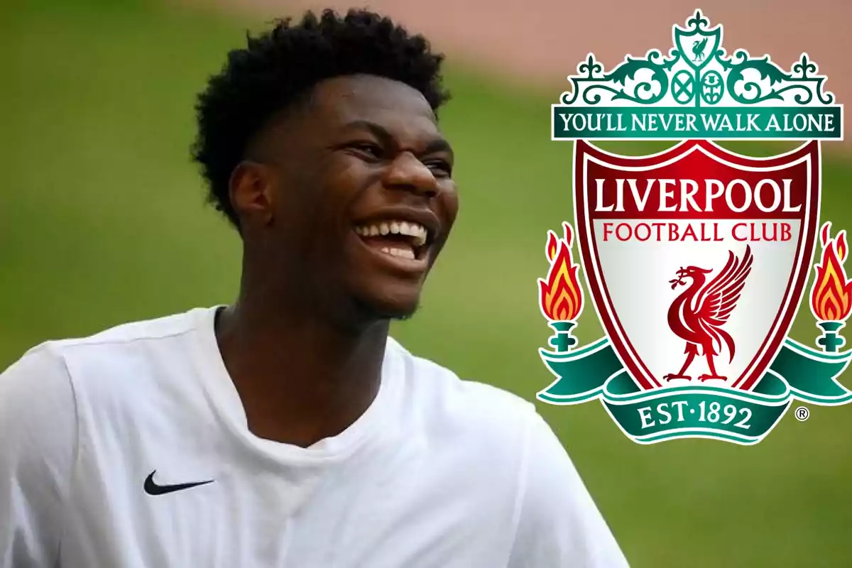 Un hombre sonriente con una camiseta blanca junto al escudo del Liverpool Football Club.