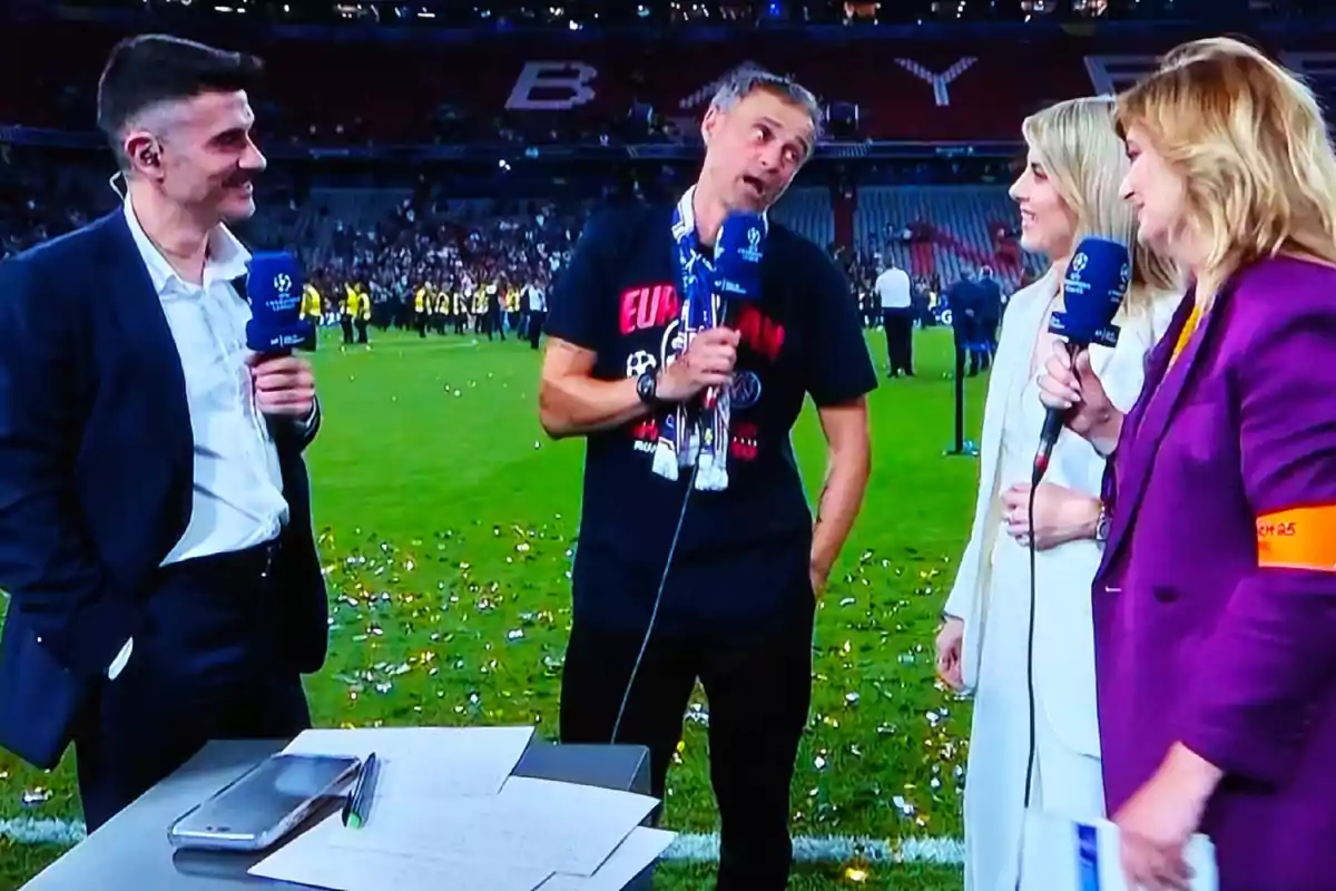 Cuatro personas están de pie en un campo de fútbol durante una entrevista en vivo, tres de ellas sostienen micrófonos y parecen estar conversando y sonriendo, mientras que al fondo se observa público y confeti en el césped. Cuatro personas están de pie en un campo de fútbol durante una entrevista en vivo, tres de ellas sostienen micrófonos y parecen estar conversando y sonriendo, mientras que al fondo se observa público y confeti en el césped.