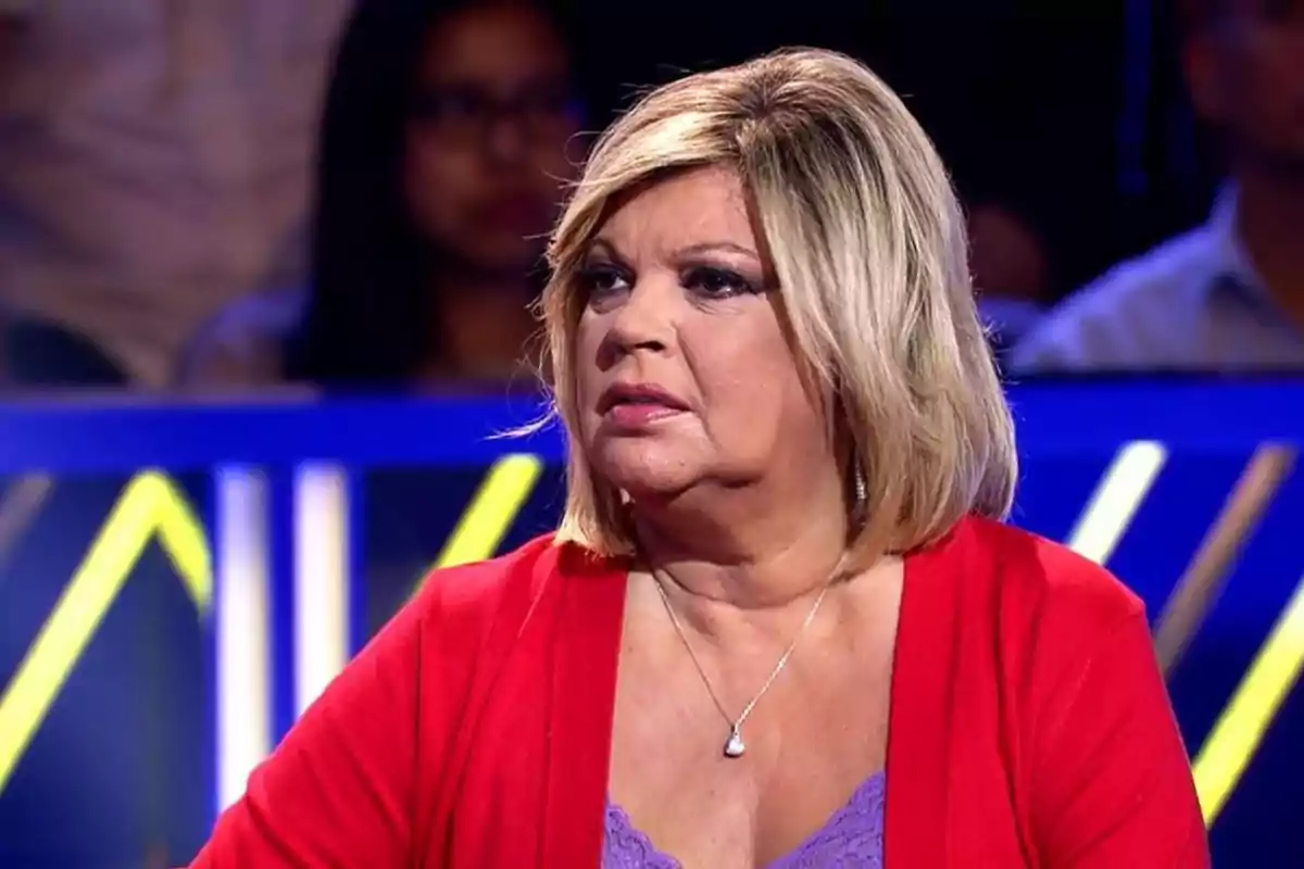 Mujer con cabello rubio y corto, usando un suéter rojo y una blusa morada, en un entorno de estudio de televisión.