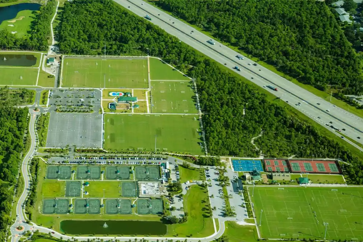 Vista aérea de un complejo deportivo con varias canchas de tenis, campos de fútbol, estacionamientos y áreas verdes junto a una autopista rodeada de árboles. Vista aérea de un complejo deportivo con varias canchas de tenis, campos de fútbol, estacionamientos y áreas verdes junto a una autopista rodeada de árboles.