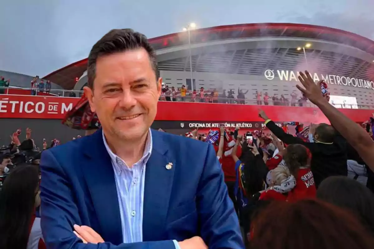 Un hombre sonriente con traje azul frente a un estadio con aficionados celebrando.