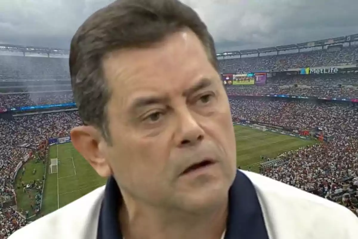 Hombre con expresión seria en primer plano con un estadio de fútbol lleno de espectadores en el fondo.