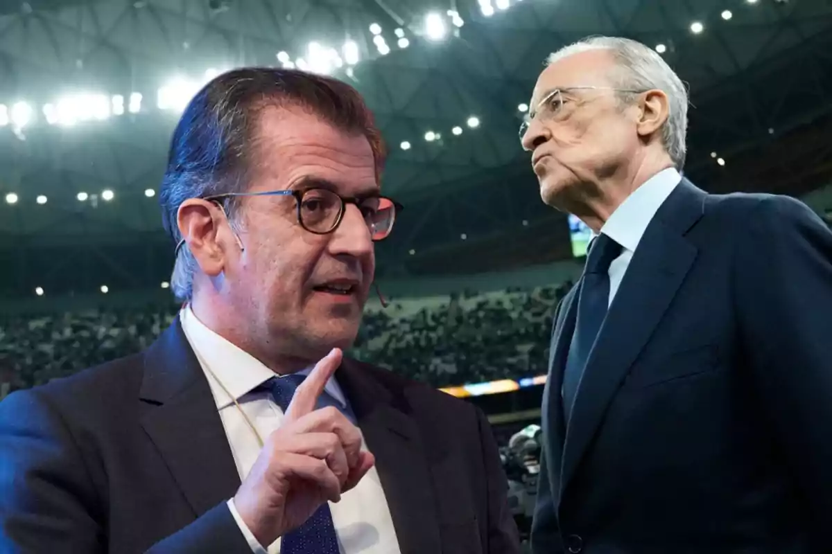 Dos hombres en traje conversando en un estadio iluminado.