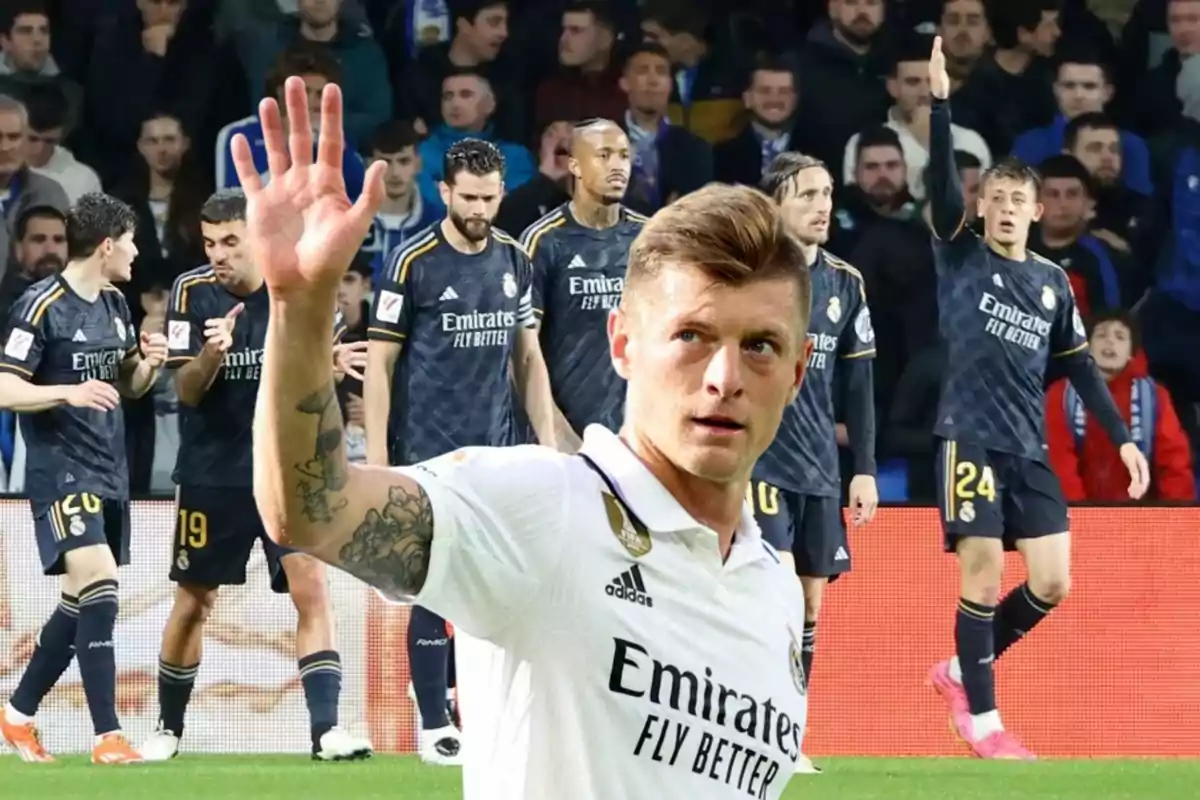 Un jugador de fútbol con la camiseta blanca del Real Madrid saluda mientras sus compañeros con la camiseta negra celebran en el fondo.