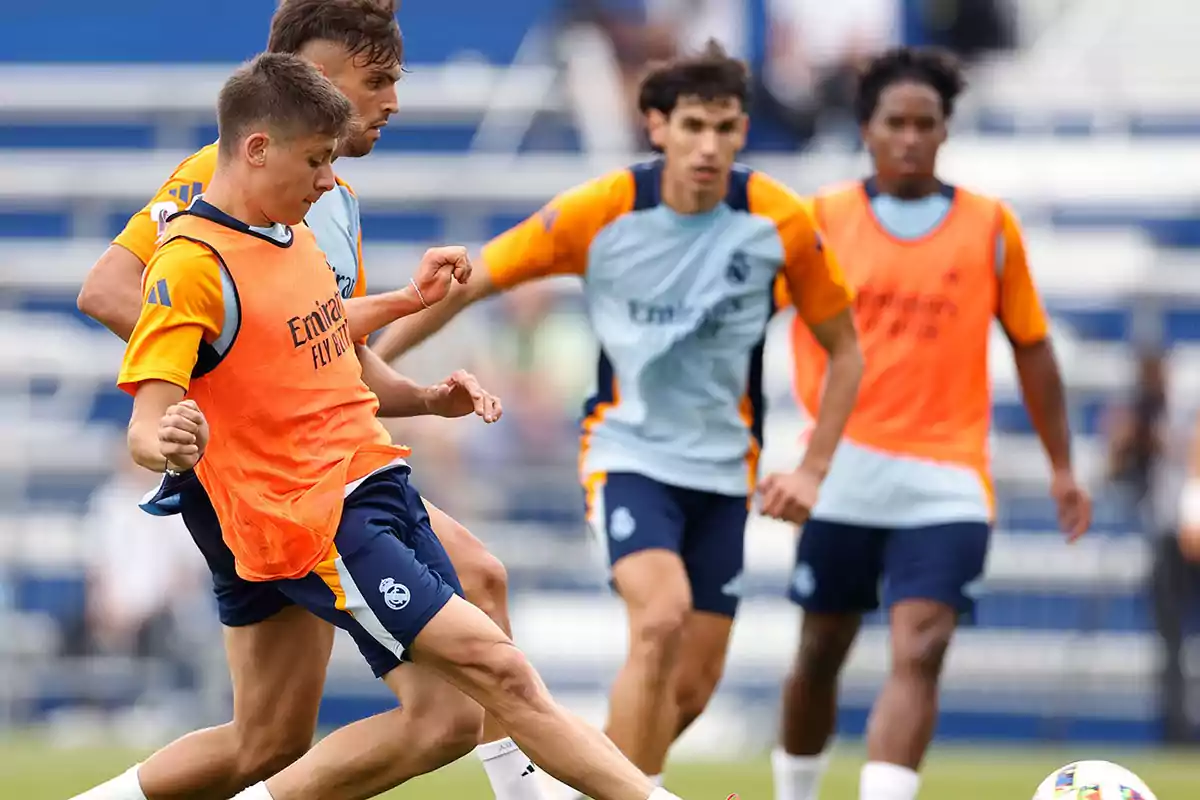 Jugadores de fútbol entrenando en un campo, algunos con petos naranjas y otros con camisetas de entrenamiento azules.