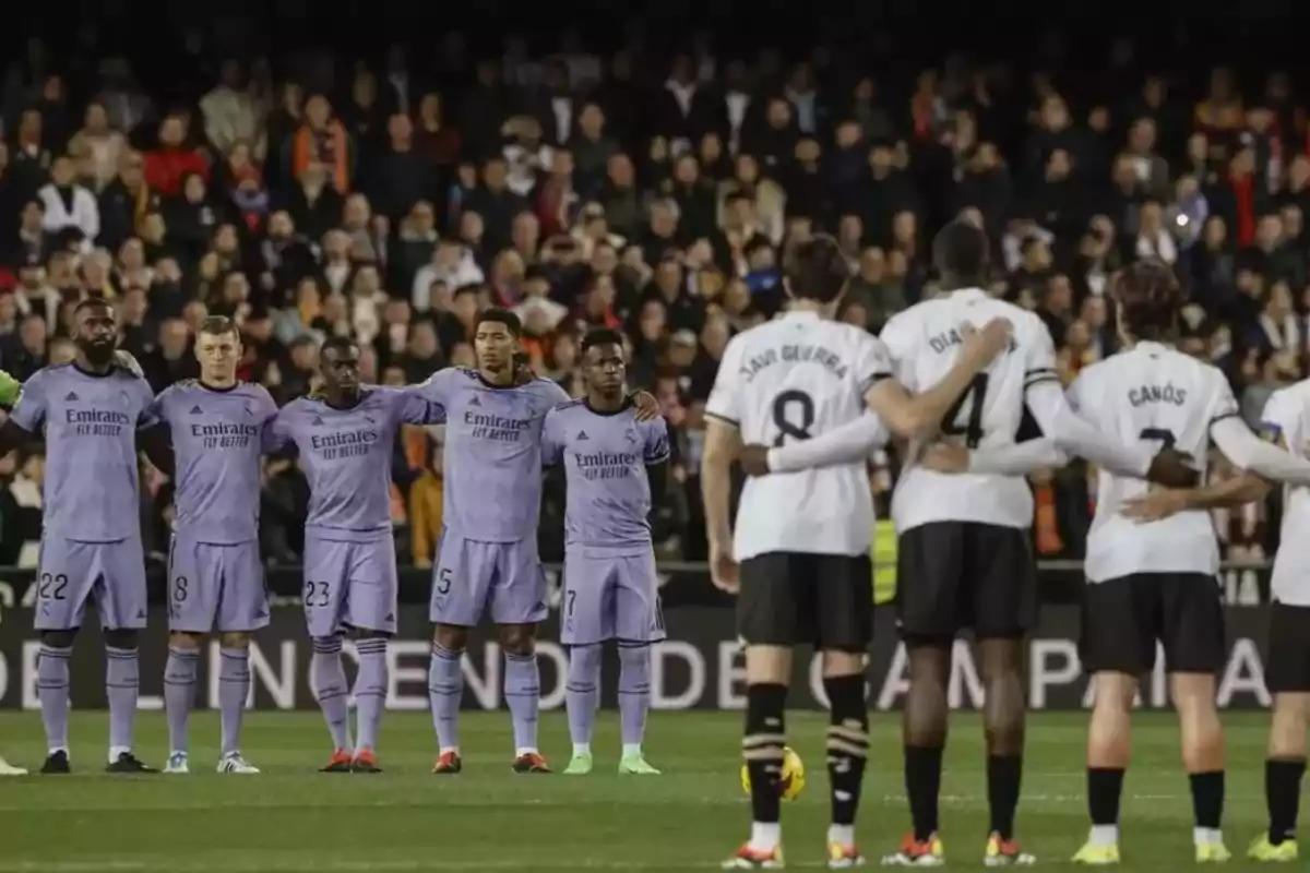 Dos equipos de fútbol se alinean en el campo antes de un partido, con jugadores abrazados en señal de respeto y unidad, mientras el público observa desde las gradas.