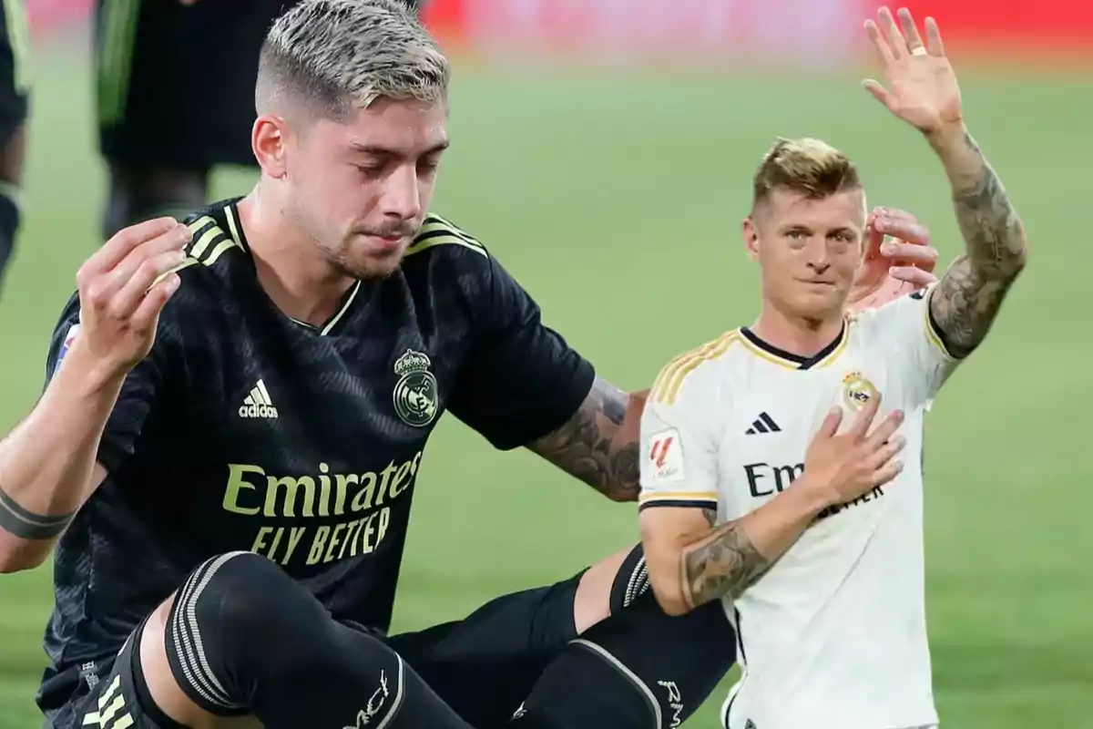 Dos jugadores de fútbol del Real Madrid en el campo, uno con uniforme negro y el otro con uniforme blanco, ambos con expresiones serias.