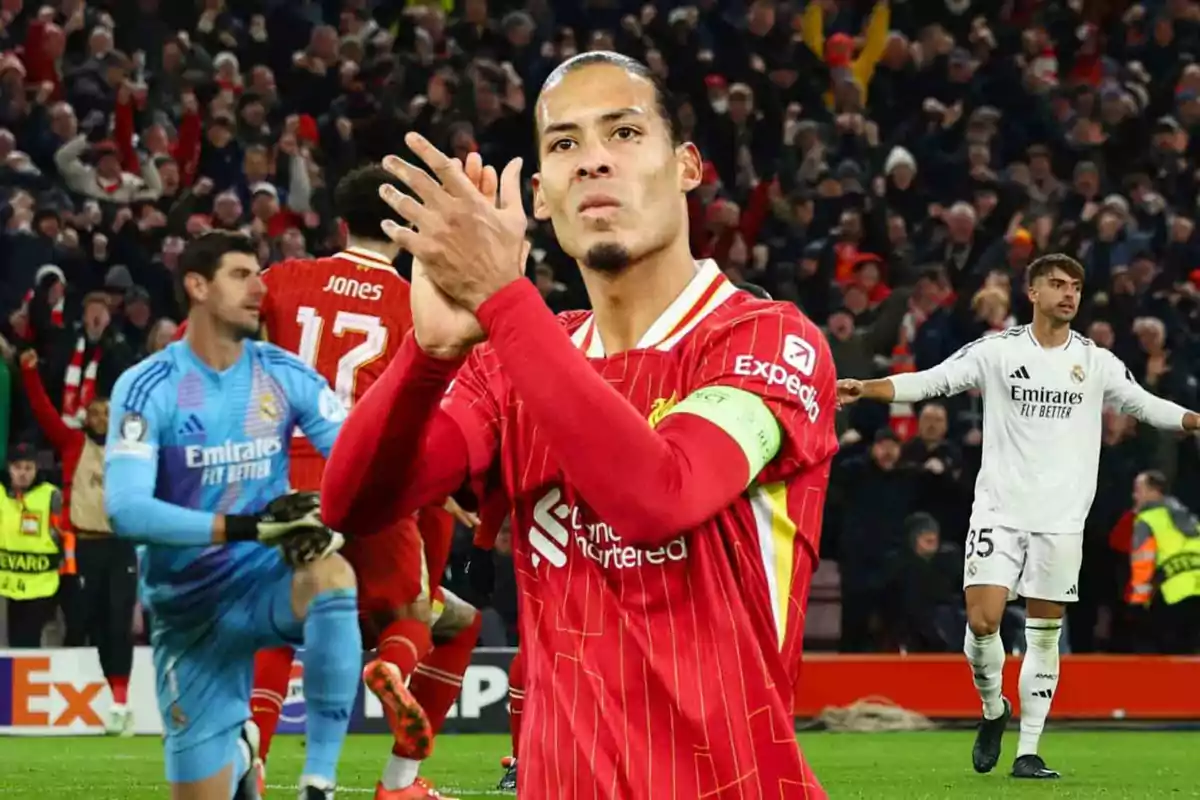 Un jugador de fútbol con uniforme rojo aplaude en el campo mientras otros jugadores y el público están al fondo.