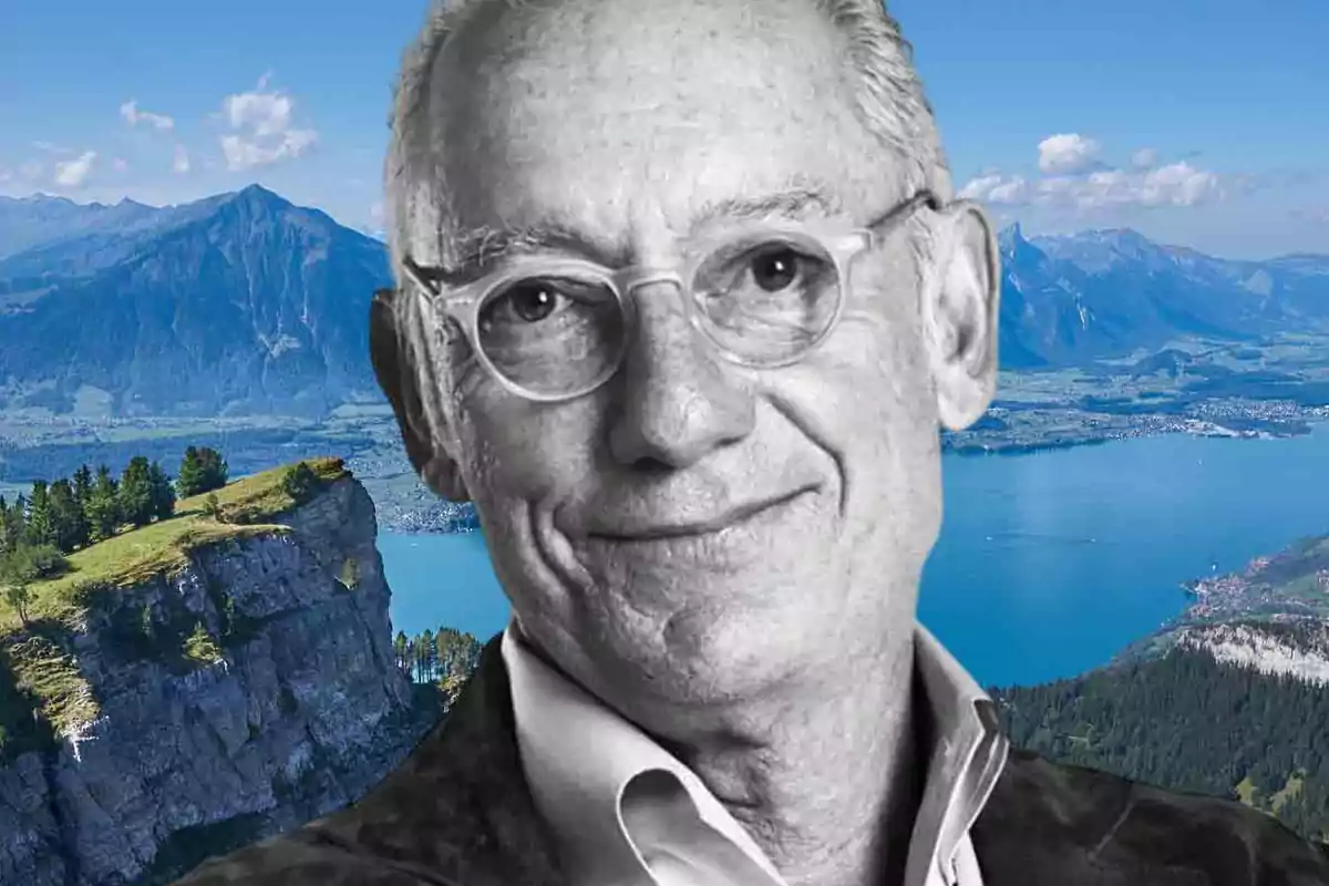 Un hombre mayor con gafas sonríe frente a un paisaje montañoso y un lago.