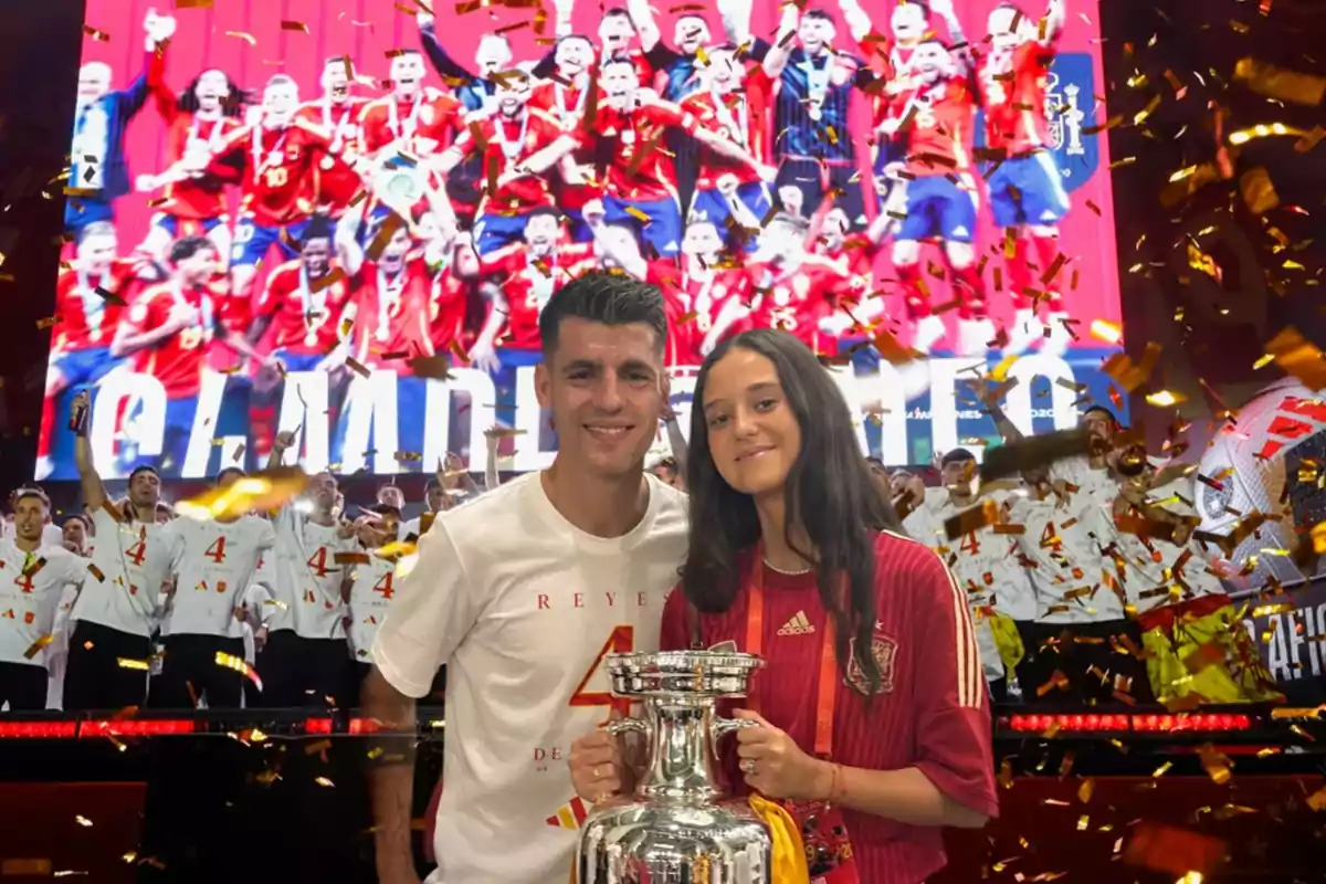 Dos personas posan con un trofeo frente a una pantalla gigante que muestra a un equipo de fútbol celebrando con confeti dorado cayendo alrededor.