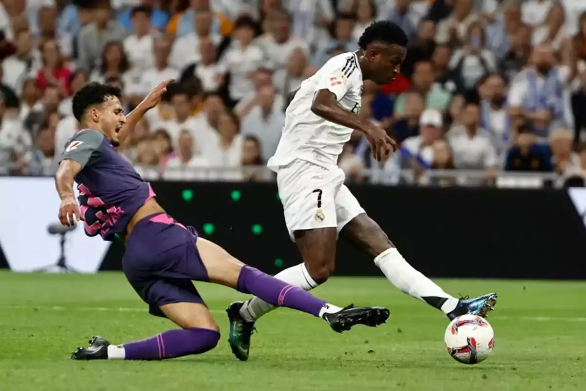 Un jugador con uniforme blanco dribla el balón mientras otro jugador con uniforme morado intenta deslizarse para quitarle el balón en un partido de fútbol.