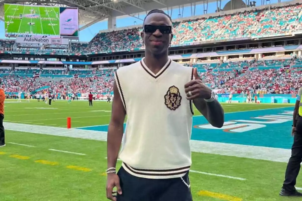 Persona con gafas de sol y chaleco blanco haciendo un gesto de aprobación en un estadio de fútbol americano. Persona con gafas de sol y chaleco blanco haciendo un gesto de aprobación en un estadio de fútbol americano.