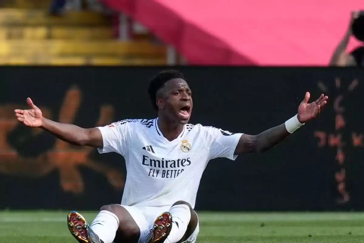 Un jugador de fútbol con uniforme blanco está sentado en el campo con los brazos extendidos y expresión de frustración. Un jugador de fútbol con uniforme blanco está sentado en el campo con los brazos extendidos y expresión de frustración.