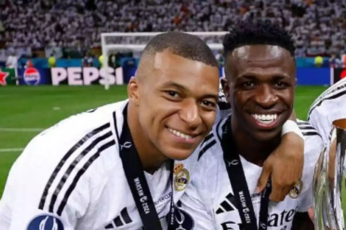 Dos futbolistas sonrientes con medallas y trofeo en un estadio.