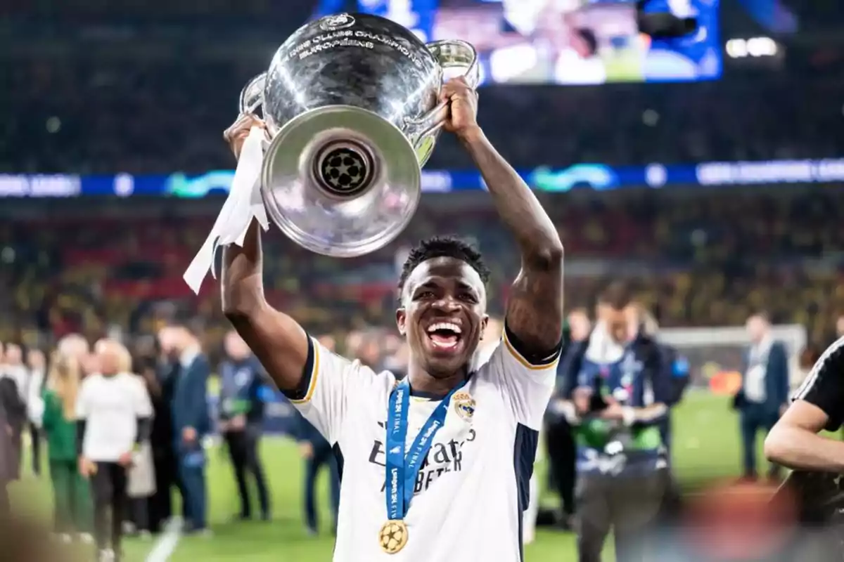 Un jugador de fútbol sonriente levanta un trofeo grande en un estadio lleno de gente.