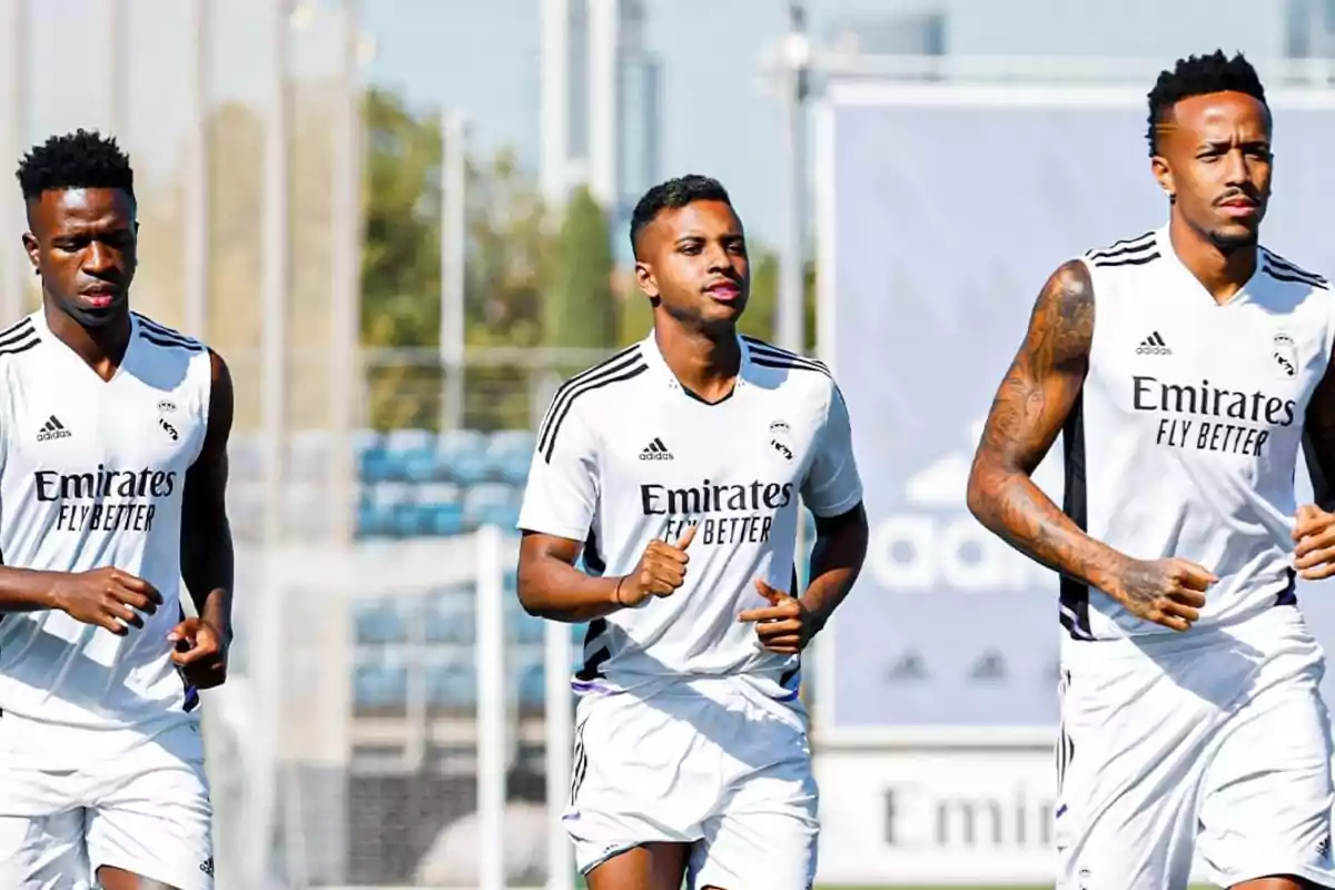 Tres jugadores de fútbol del Real Madrid corriendo durante un entrenamiento, vistiendo uniformes blancos con el logo de Emirates Fly Better.