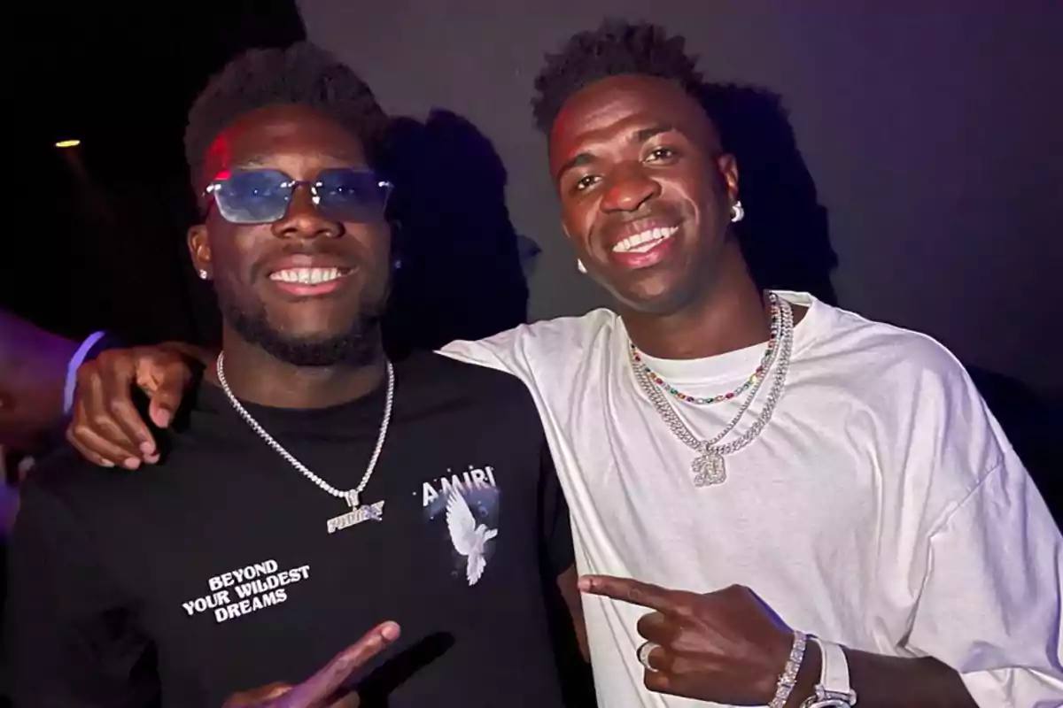 Dos personas sonrientes posan juntas en un ambiente oscuro, una lleva gafas de sol y una camiseta negra, la otra viste una camiseta blanca y collares.