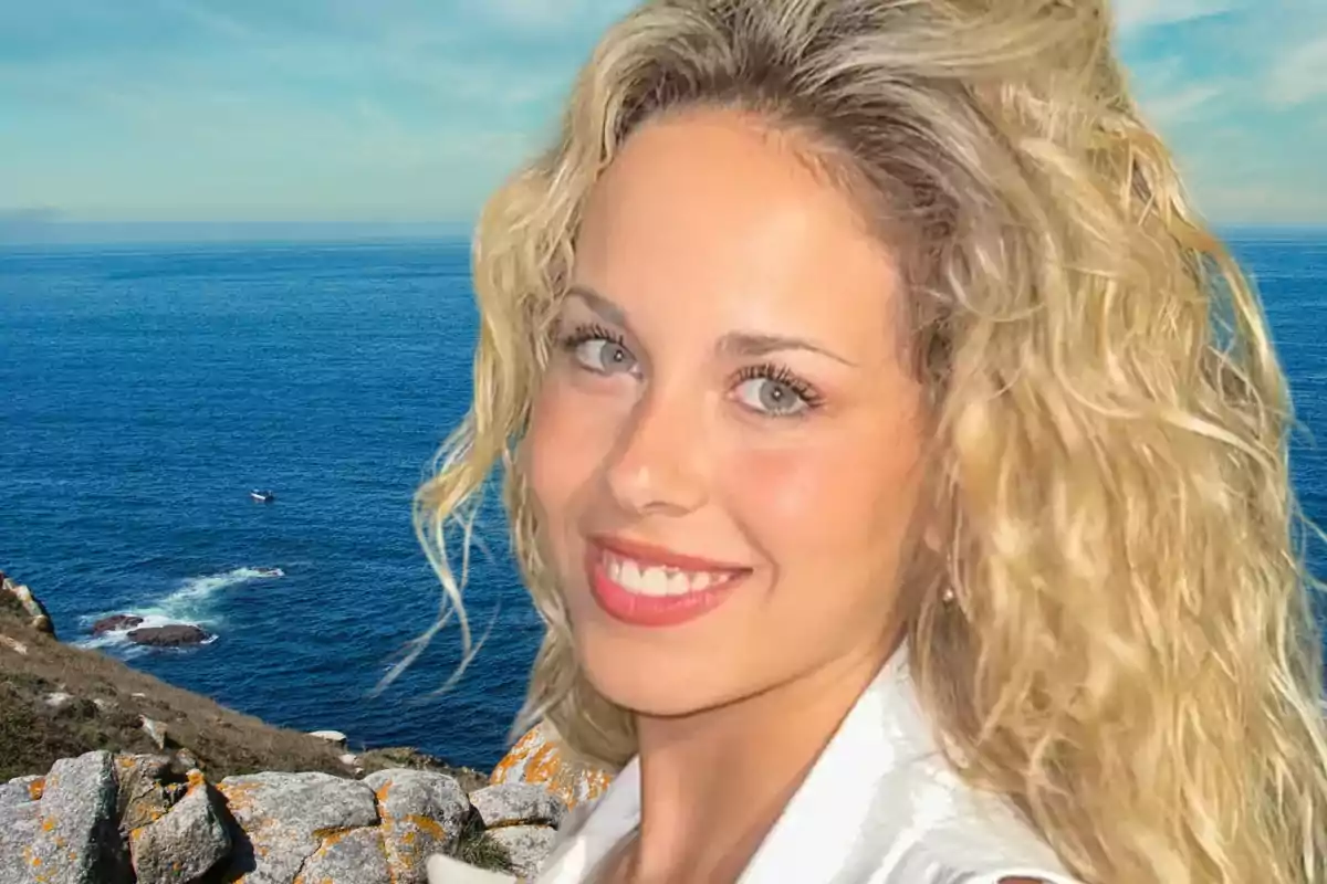 Una mujer sonriente con cabello rubio rizado frente a un paisaje costero con mar azul y rocas.