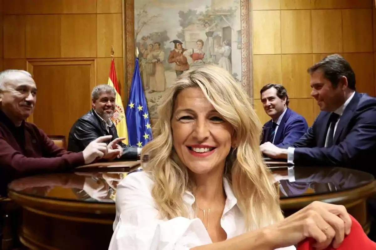 Una mujer sonriente en primer plano con cuatro hombres sentados alrededor de una mesa redonda en una sala de reuniones con banderas de España y la Unión Europea en el fondo.