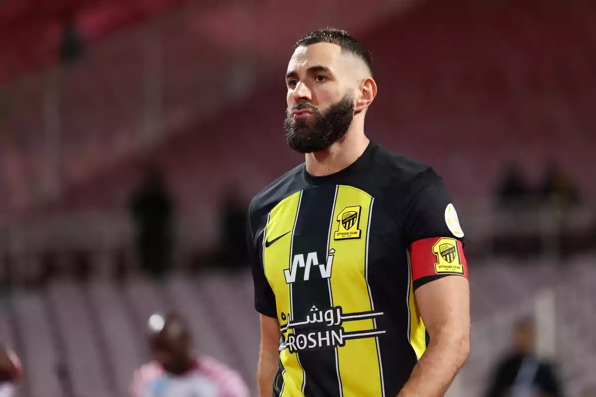 Un jugador de fútbol con barba y cabello corto viste el uniforme del Al-Ittihad Club, con una camiseta de rayas amarillas y negras y un brazalete de capitán rojo, en un estadio.