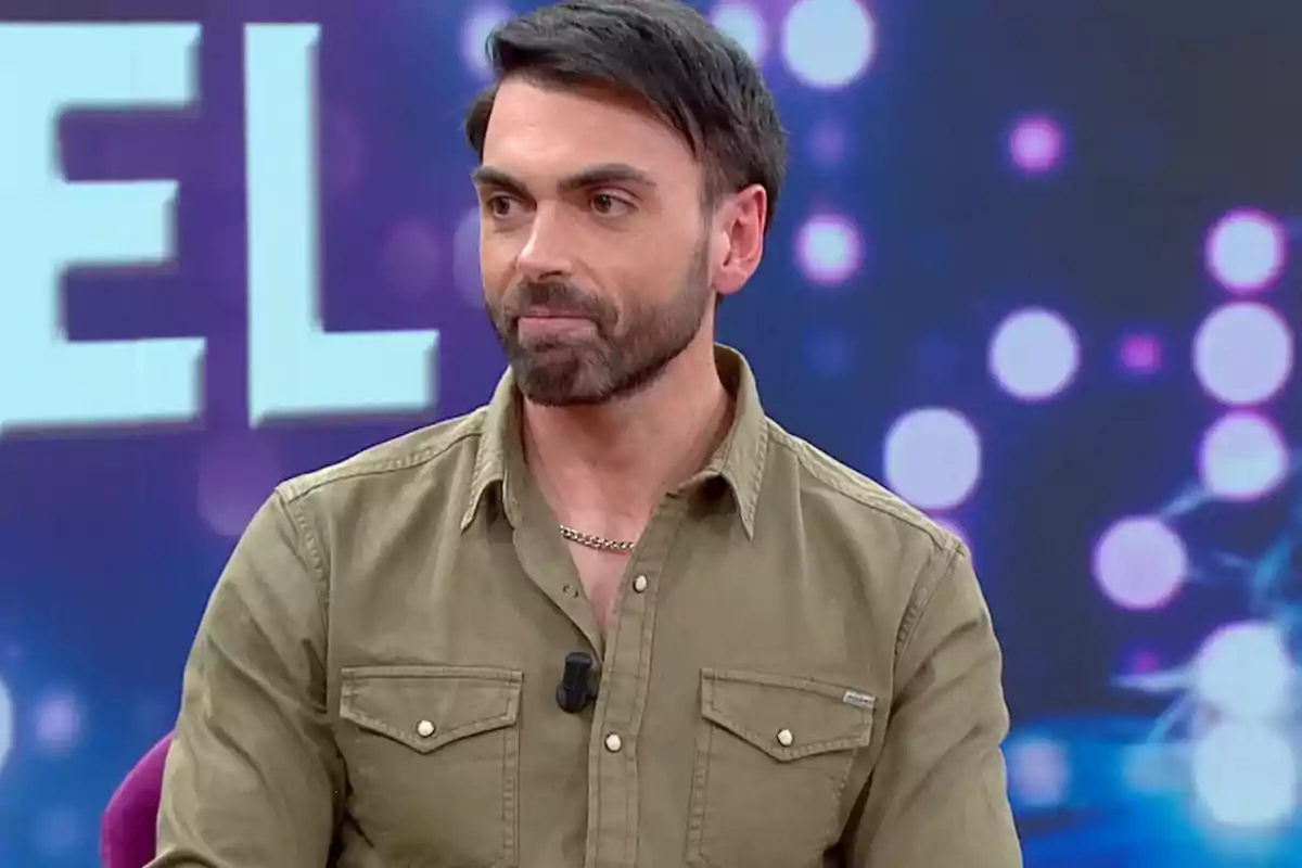 Un hombre con barba y camisa verde aparece en un programa de televisión con un fondo de luces desenfocadas.